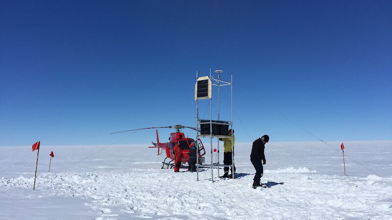 Scientists installing equipment on the Totten Glacier with a red helicopter in the background.