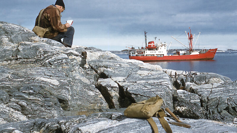 A scientist sits on a rock overlooking a bay writing in his field notebook.