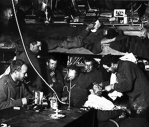 Frank Hurley trims John Hunter’s beard in the tight confines of the Main Hut at Cape Denison, with five of their fellow expeditioners looking on