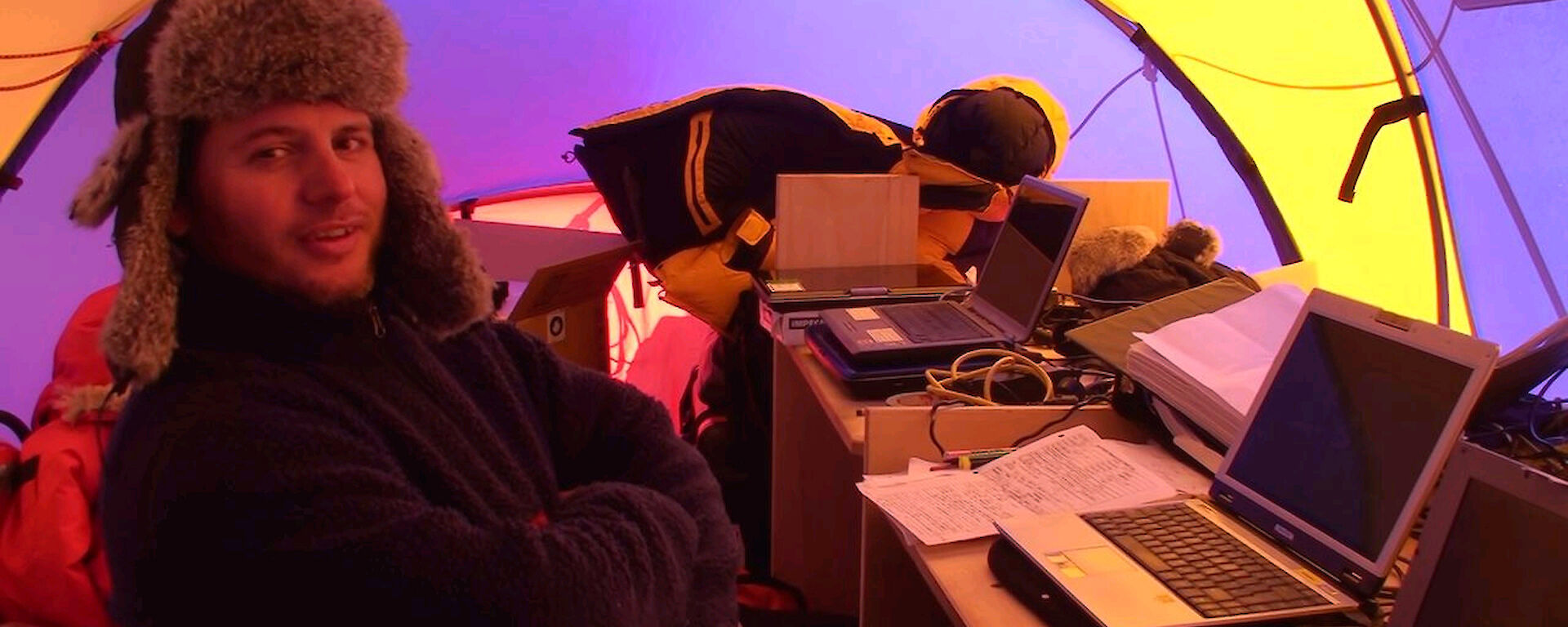 Scientist in furred hat, at desk in science tent, with laptops and equipment