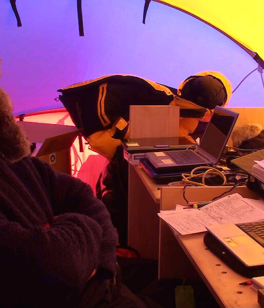 Scientist in furred hat, at desk in science tent, with laptops and equipment