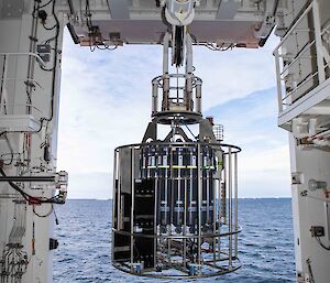 A CTD rosette being hoisted by an overhead crane to deploy through an opening in the side of the ship. Open ocean, ice and cloudy skies in background.
