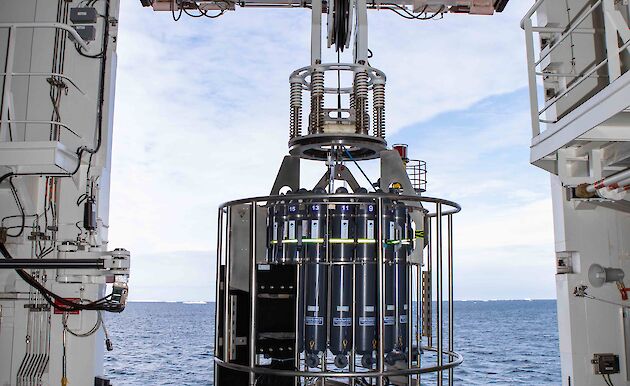 A CTD rosette being hoisted by an overhead crane to deploy through an opening in the side of the ship. Open ocean, ice and cloudy skies in background.