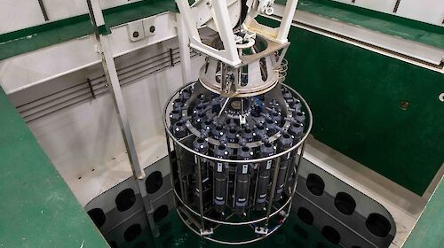 A large cylinder containing many capsules is lowered into a pool.