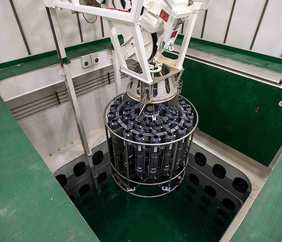 A large cylinder containing many capsules is lowered into a pool.