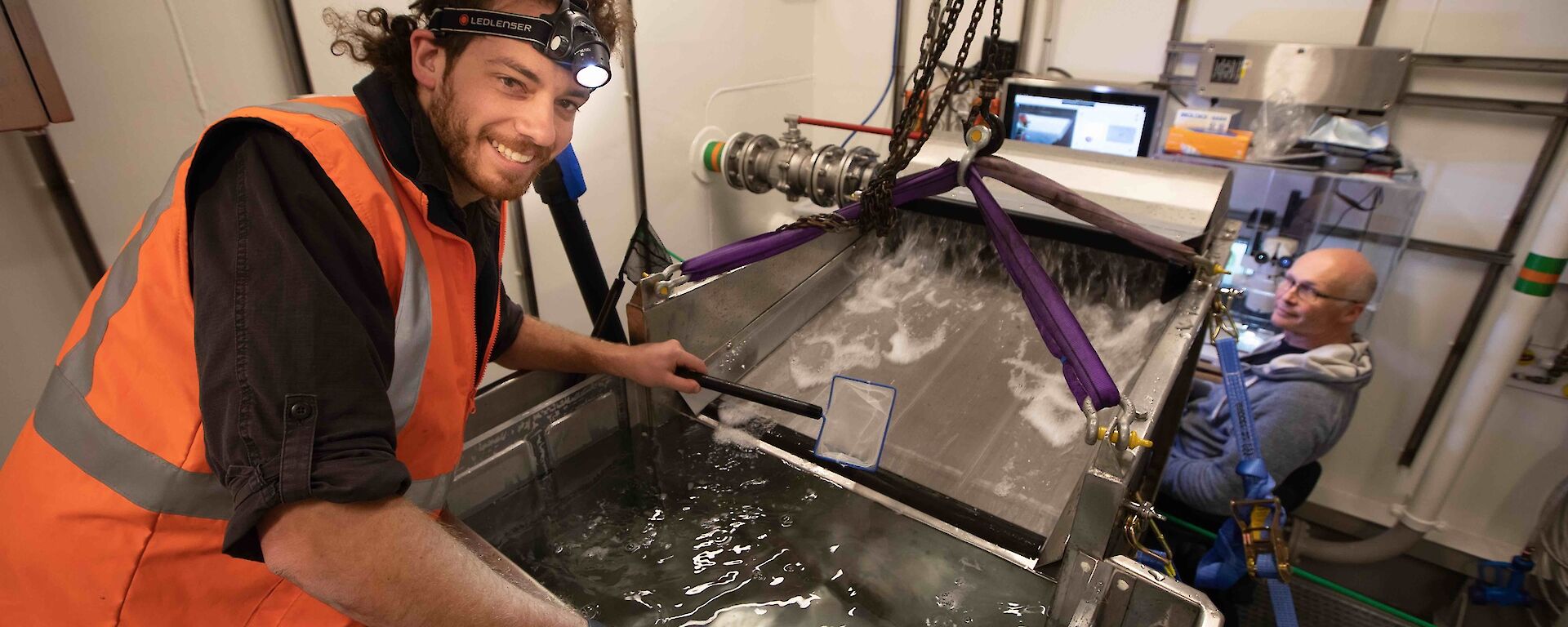 A scientist leans over a tank of water, surrounded by equipment. Another scientist sits as a computer. Both are smiling.