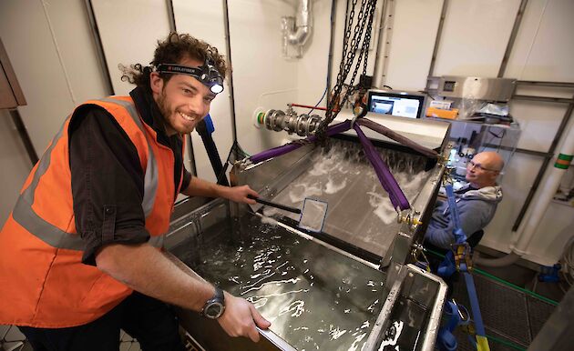 A scientist leans over a tank of water, surrounded by equipment. Another scientist sits as a computer. Both are smiling.