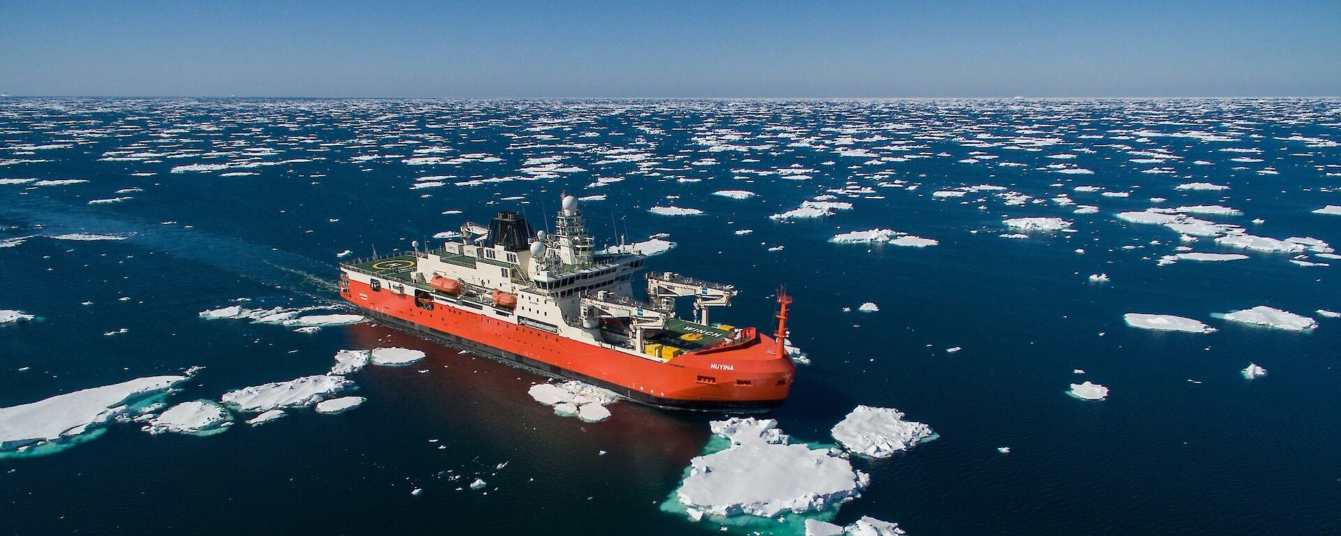 An orange and white ship sails through the ocean, surrounded by floating chunks of ice. The sky is clear and blue.