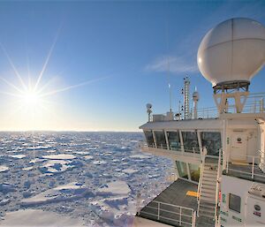 Looking back along starboard side of icebreaker pushing through sea ice with sun glaring in blue sky.