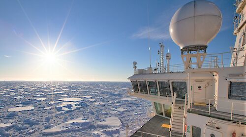 Looking back along starboard side of icebreaker pushing through sea ice with sun glaring in blue sky.