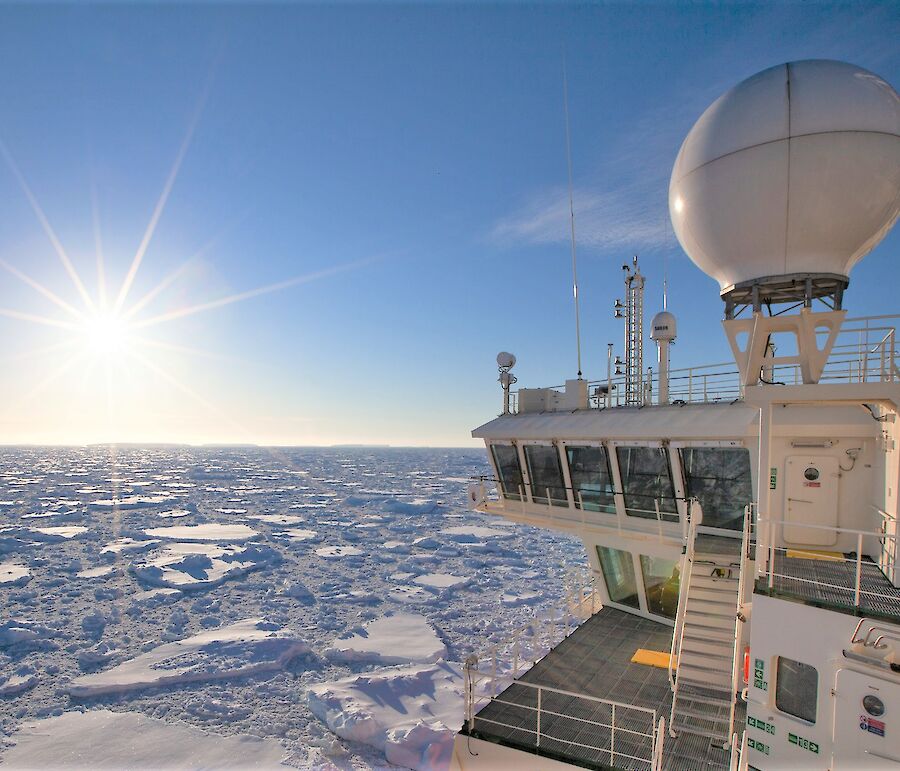 Looking back along starboard side of icebreaker pushing through sea ice with sun glaring in blue sky.