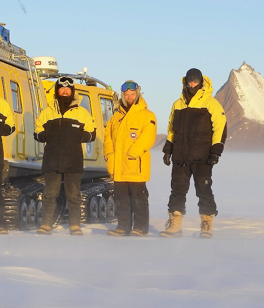 Expeditioners stand in front of a yellow Hägglunds in a snowy landscape.