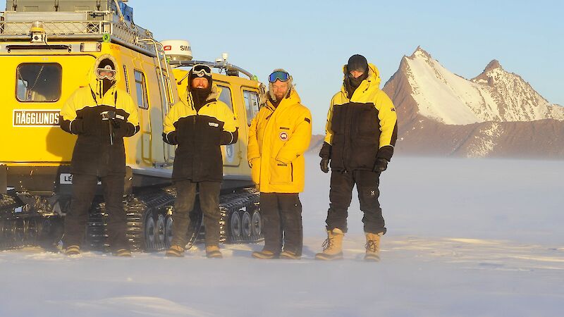 Expeditioners stand in front of a yellow Hägglunds in a snowy landscape.