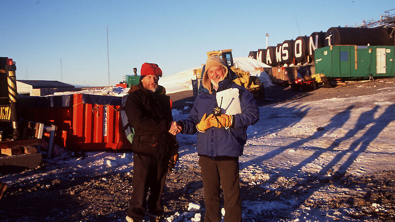 Chief scientist farewells Mawson station leader outdoors with station infrastructure behind