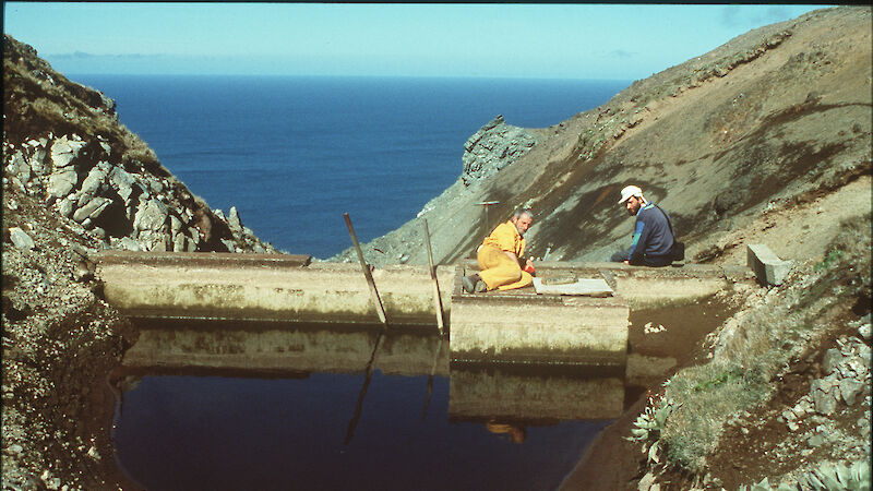 Two expeditioners sitting on small dam with bay in background