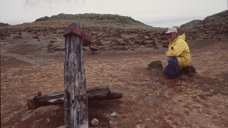 Man squatting beside grey weathered baulks of timber and iron banding remains of wireless mast