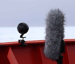 A fluffy microphone and a spherical camera sit on the deck of an orange ship.