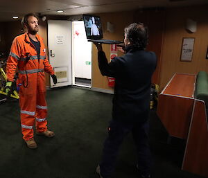 An artist holds a laptop up in front of a crew member dressed in orange in a dark room. On the laptop screen, there is an image of the crew member.