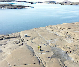 Aerial photo looking down at 2 expeditioners standing on rocky ground with water behind them.