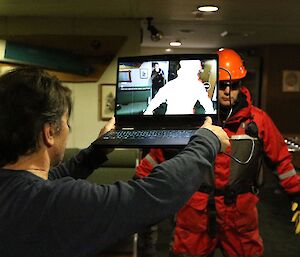 An artist holds a laptop up in front of a crew member dressed in orange in a dark room. On the laptop screen, there is an image of the crew member.