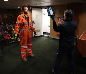 An artist holds a laptop up in front of a crew member dressed in orange in a dark room. On the laptop screen, there is an image of the crew member.