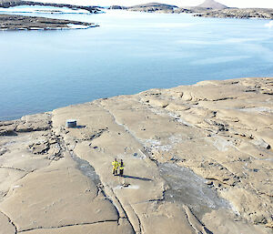 Aerial photo looking down at 2 expeditioners standing on rocky ground with water behind them.