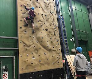 A person wearing a harness and helmet climbing the indoor rock climbing wall, with another person standing at the bottom watching.