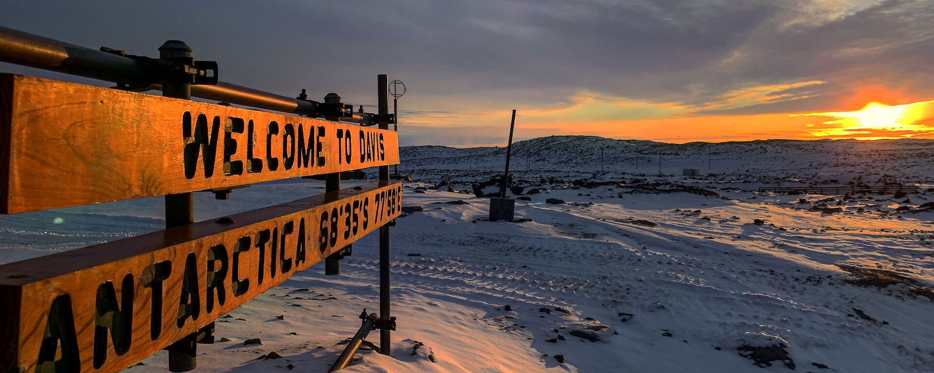 The sun sits low and yellow in the sky, illuminating a snowy landscape. a sign in the foreground says "Welcome to Davis Antarctica"