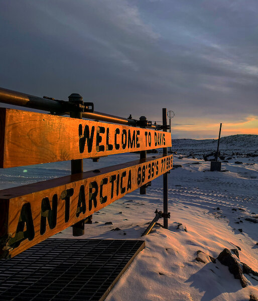 The sun sits low and yellow in the sky, illuminating a snowy landscape. a sign in the foreground says "Welcome to Davis Antarctica"