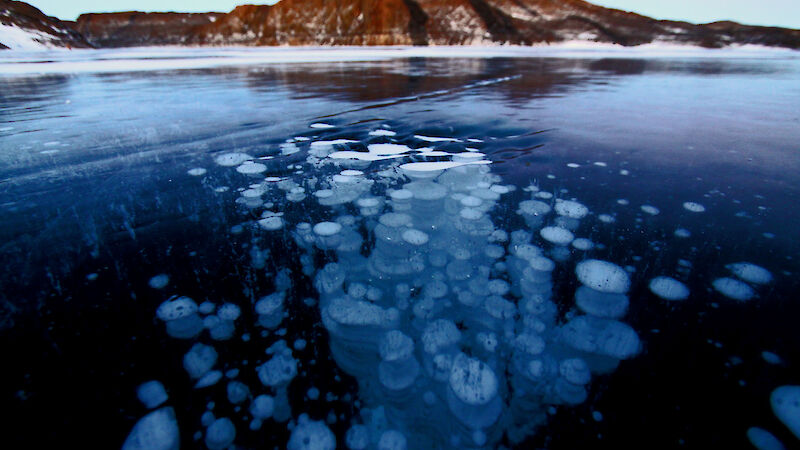 Trapped bubbles in a frozen lake are in the foreground of a landscape shot with small mountain in background