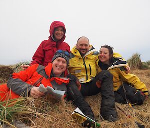 A mixed group of expeditioners pose sitting on tussock grass while smiling.