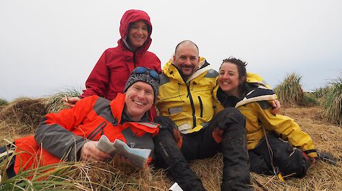 A mixed group of expeditioners pose sitting on tussock grass while smiling.