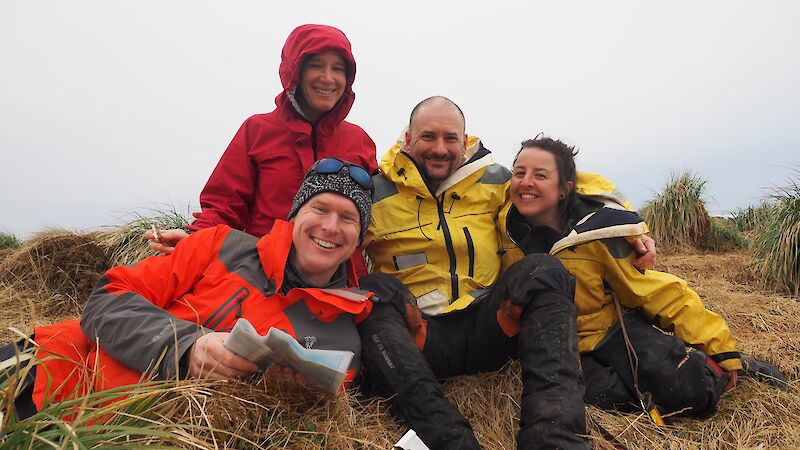 A mixed group of expeditioners pose sitting on tussock grass while smiling.
