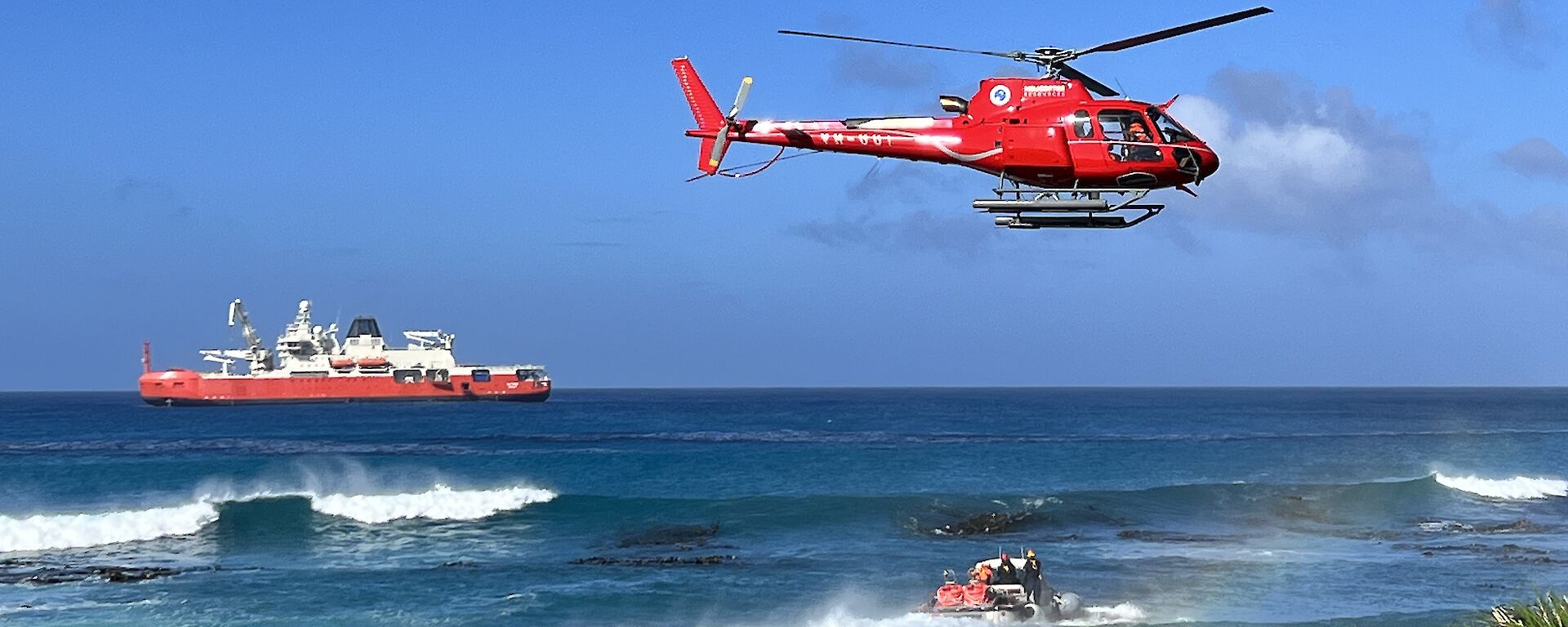 An amphibious vehicle motors through the ocean towards an orange and white ship. A red helicopter flies above.