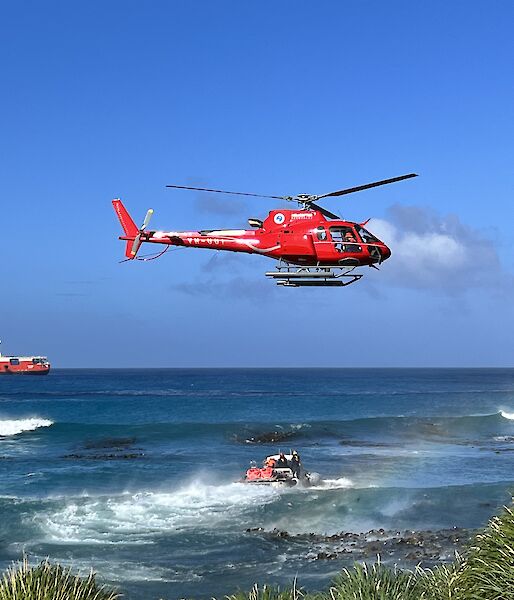 An amphibious vehicle motors through the ocean towards an orange and white ship. A red helicopter flies above.