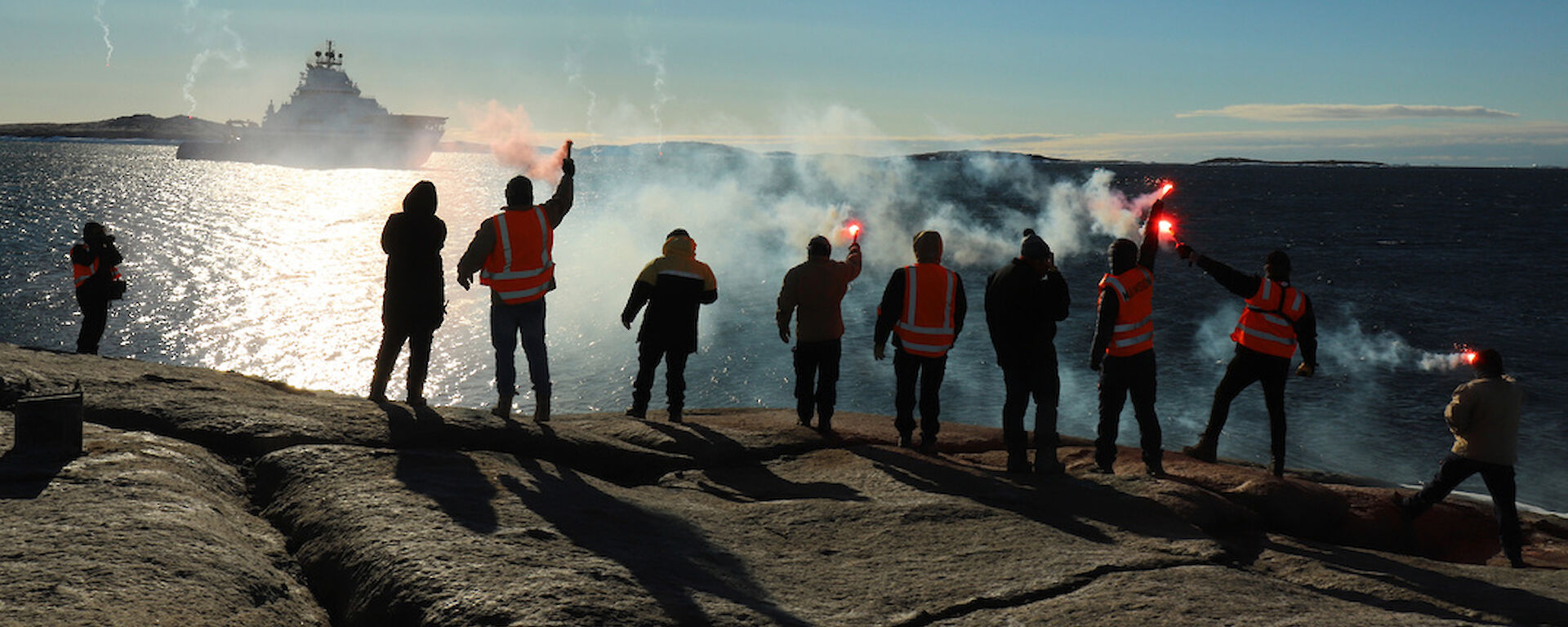 Ten people standing in a line on a rocky outcrop, with backs to camera and holding lit flares. They wave farewell to a ship in the distance.