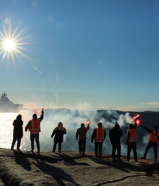 Ten people standing in a line on a rocky outcrop, with backs to camera and holding lit flares. They wave farewell to a ship in the distance.