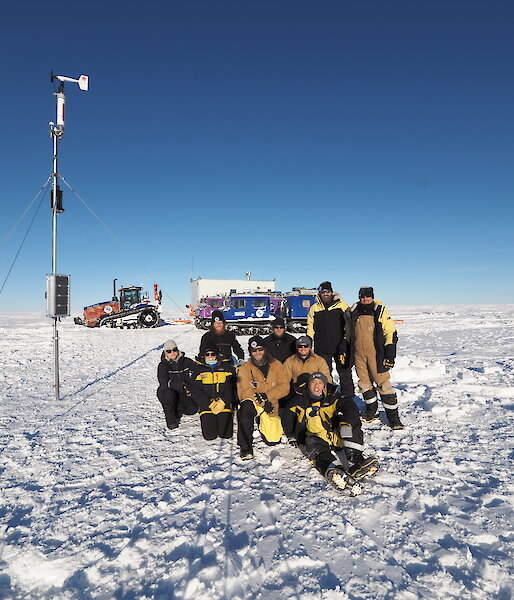 A group of expeditioners pose in an icy landscape with colourful vehicles in the distance.