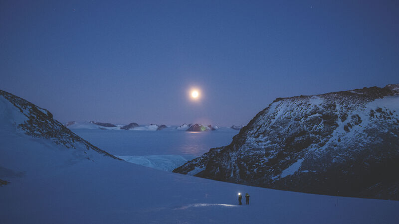 A snowy mountainous landscape bathed in a dark blue hue. The moon shines above and two people can be seen silhouetted in the distance with torches on.