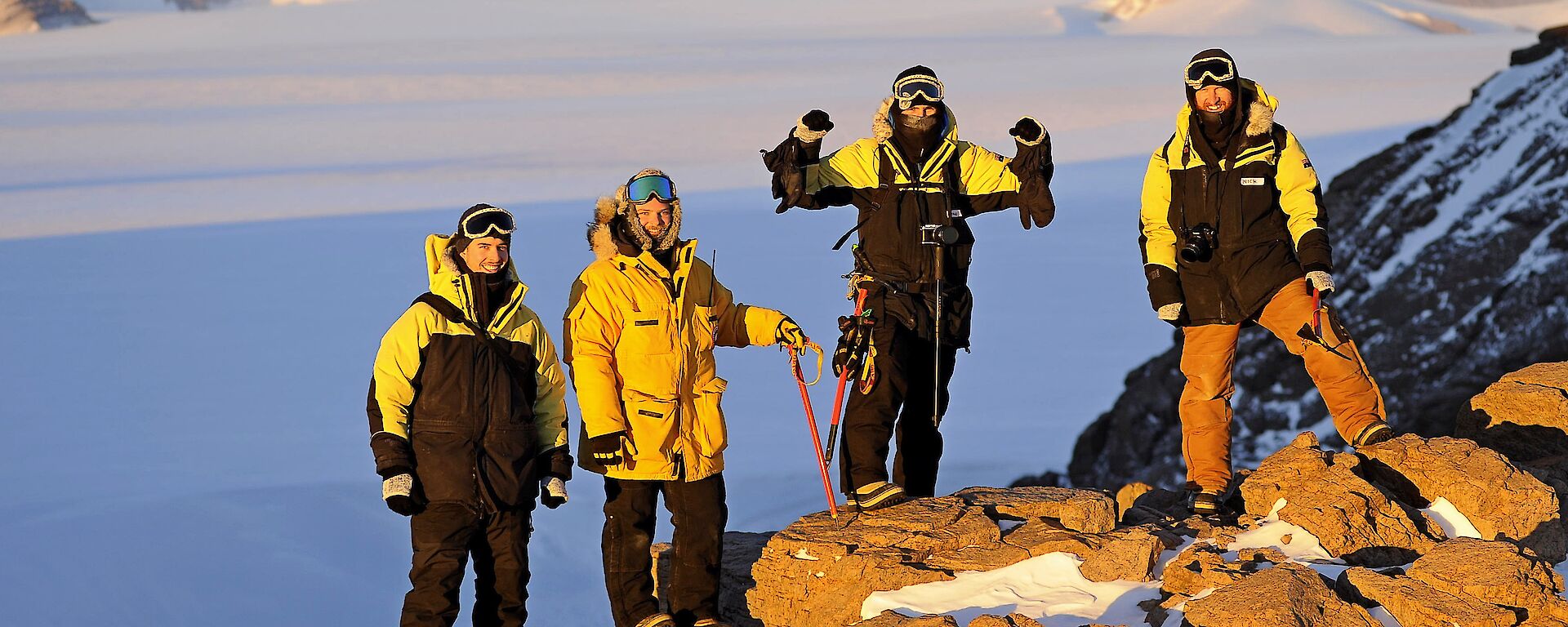 Four expeditioners stand on a rocky mountain surface with the ice plateau behind
