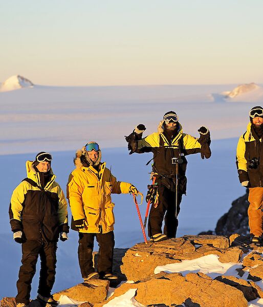 Four expeditioners stand on a rocky mountain surface with the ice plateau behind