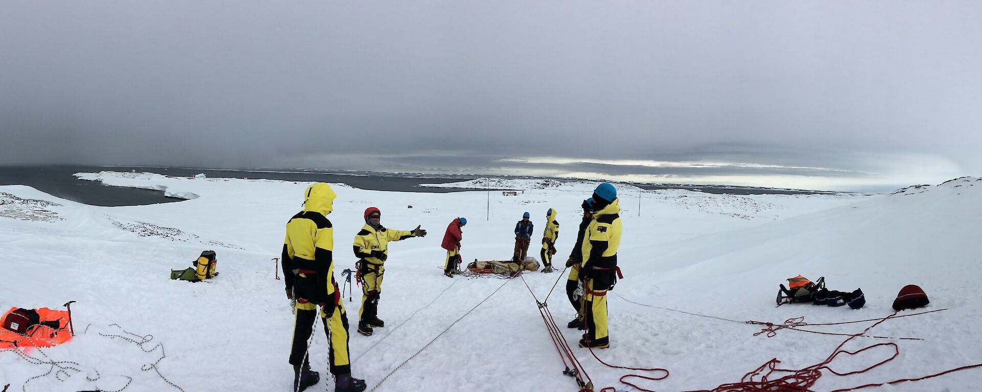 Wide angle view of stretcher connected to rope system with expos on either side listening to instructor. Set up on snowy slope with sea and dark rain clouds in background