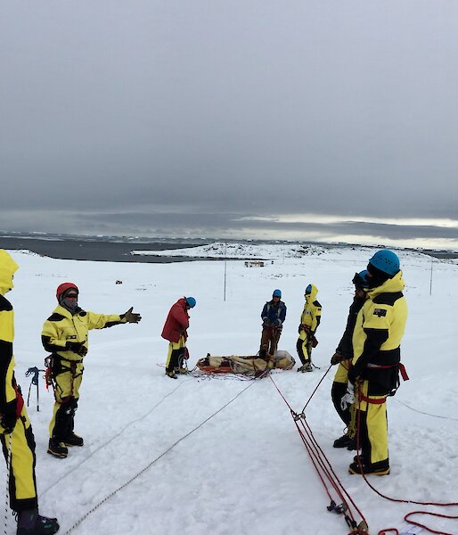 Wide angle view of stretcher connected to rope system with expos on either side listening to instructor. Set up on snowy slope with sea and dark rain clouds in background