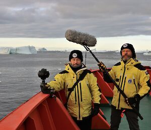 two artists with recording equipment on icebreaker with icebergs in background