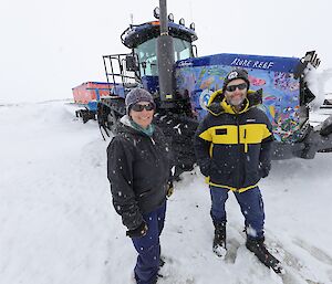 2 expeditioners stand in front of a tractor in the snow.