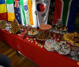 A long table draped in red cloth is covered in various, delicious looking desserts on Christmas Day dinner. Flags from various nations adorn the wall behind.