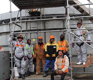 A group of seven workers stand in front of a grey water tank that they have been cleaning.