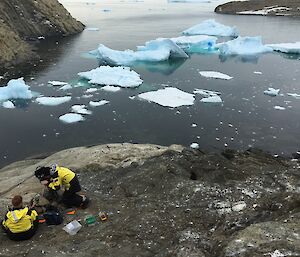 Two expeditioners maintain a camera on the edge of the island. Blue ice fragments float in the background.