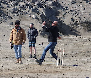 A man bowls a ball down the cricket pitch while two other men look on from the field