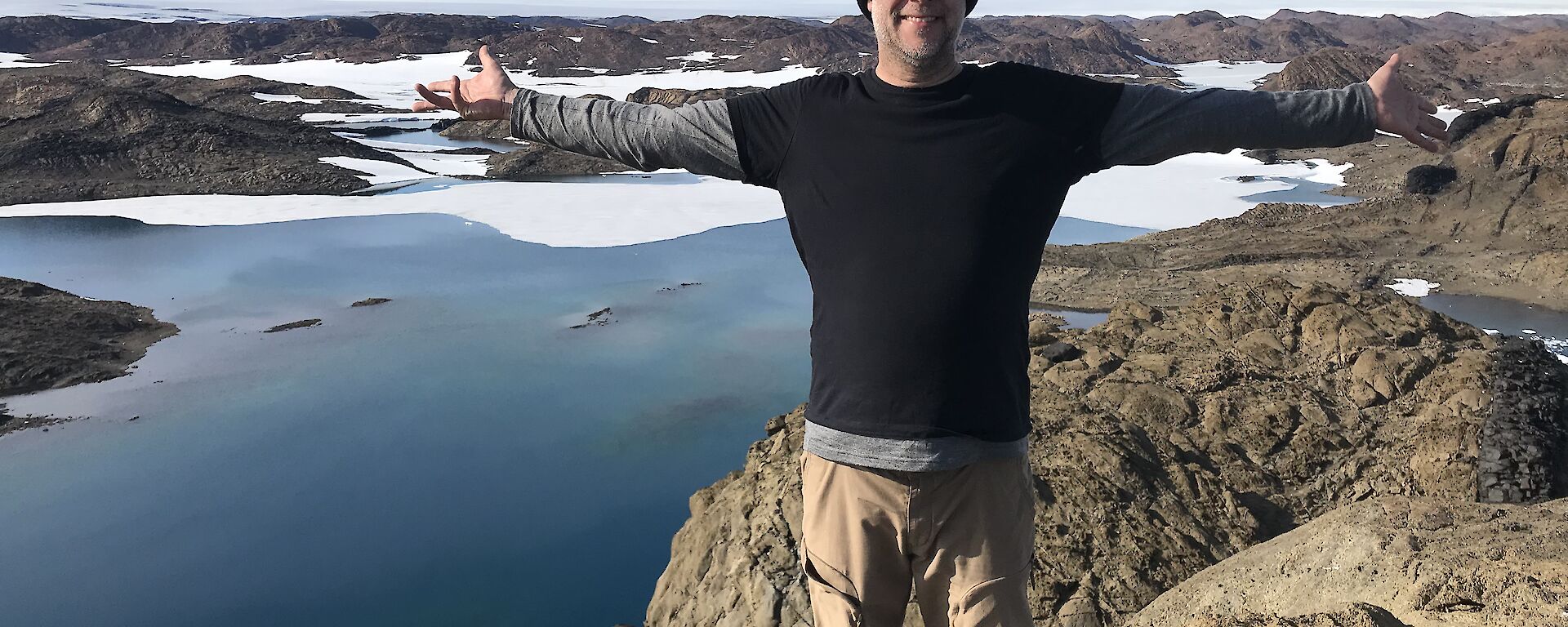 Expeditioner stands on a peak high above a vast blue lake surrounded by brown rocky hills